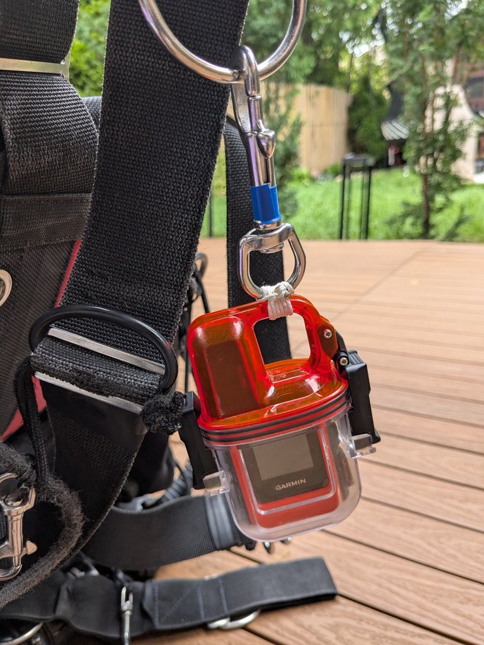 A Garmin inReach Mini in its clear protective dive case, clipped by a carabiner to a D-ring on a BCD harness.