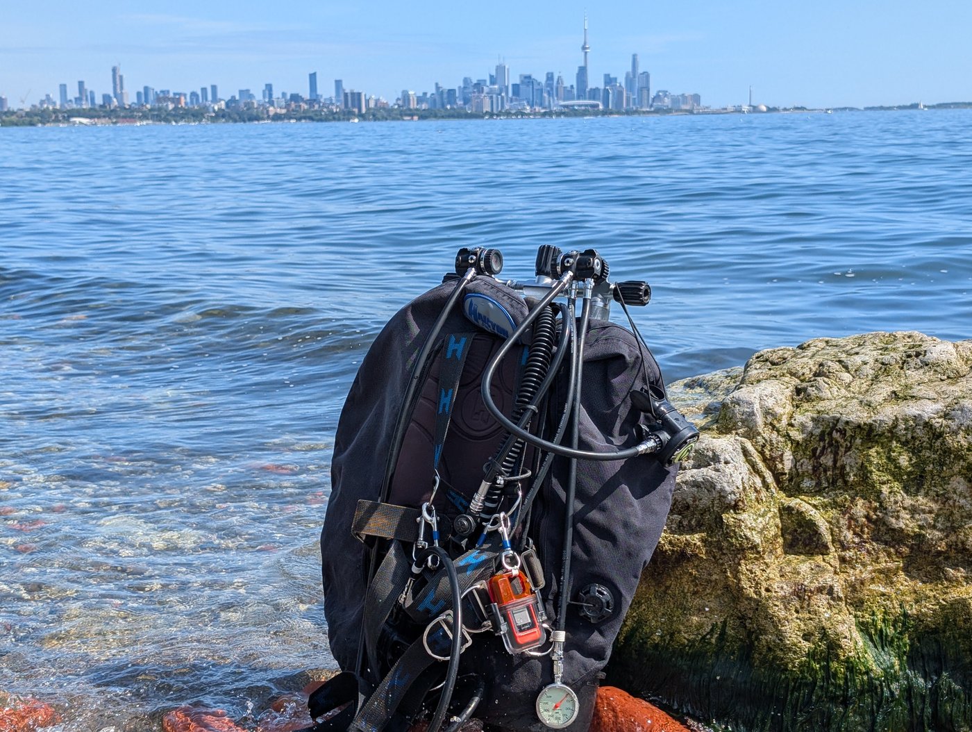 A twin-tank scuba rig staged on rocks at a Lake Ontario shore dive site, with the Toronto skyline visible across the water.